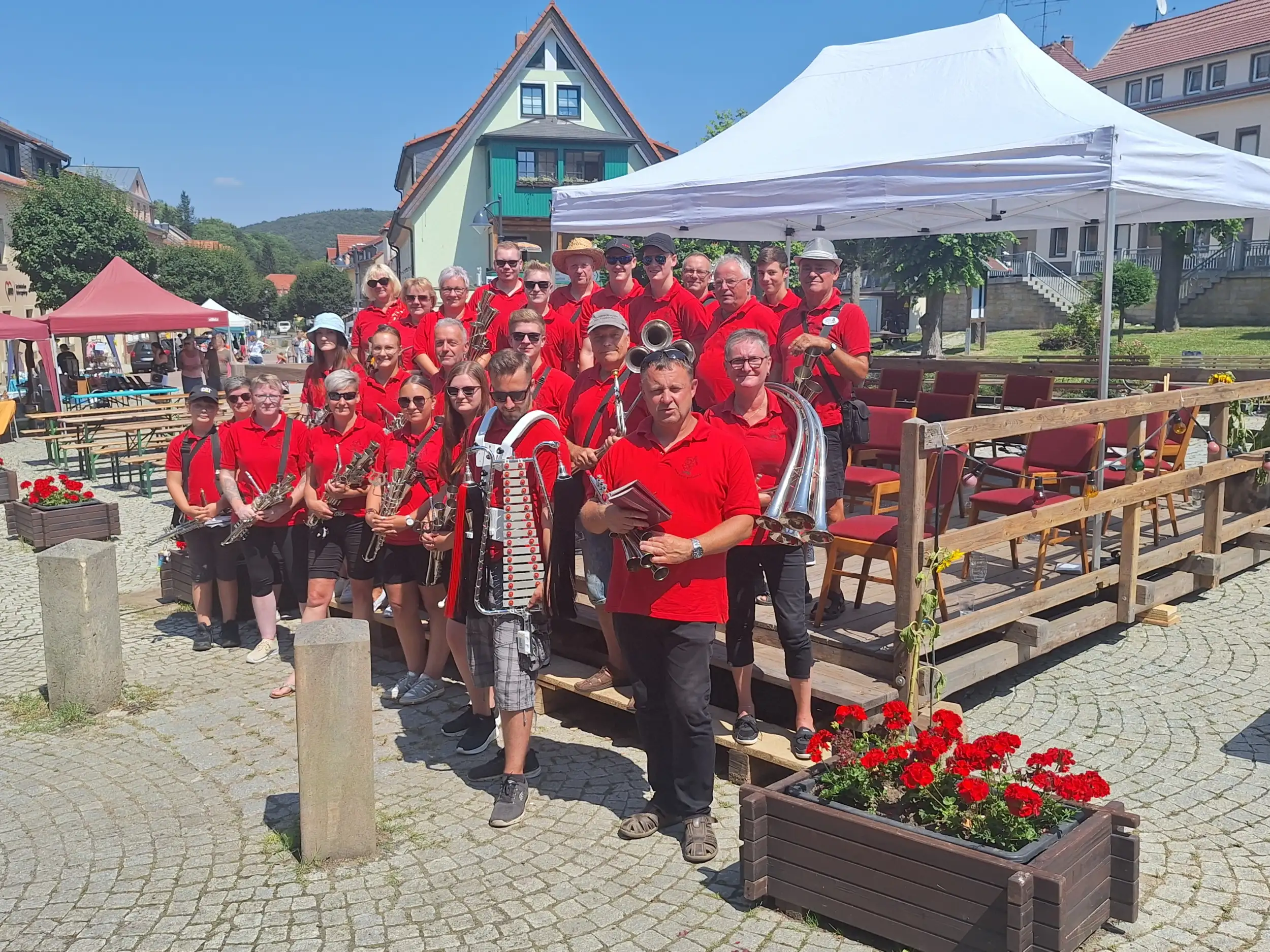 Musiker der Schalmeienkapelle Bad Gottleuba stehen in der Gruppe vor einem Pavillon auf einem gepflasterten Platz, der von Häusern umgeben ist.