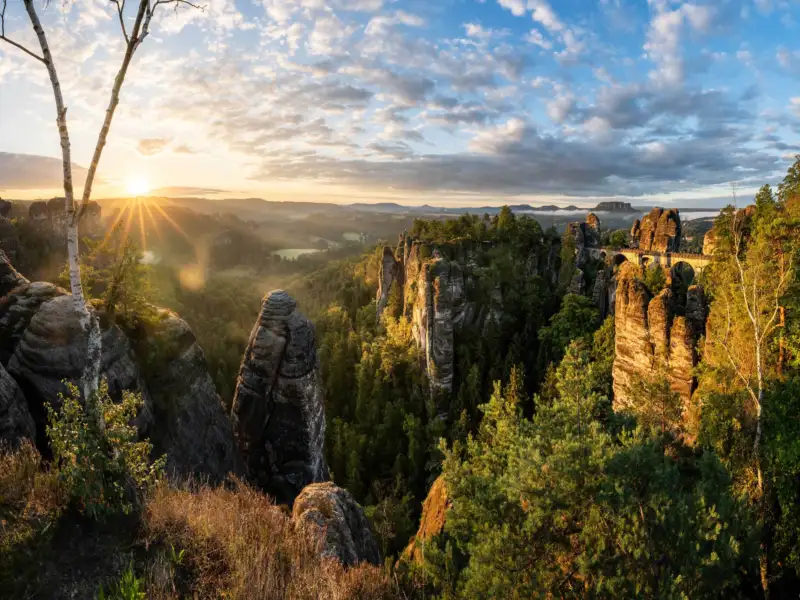 Rock formations and a bridge in a green landscape at sunrise, with a clear sky and scattered clouds.