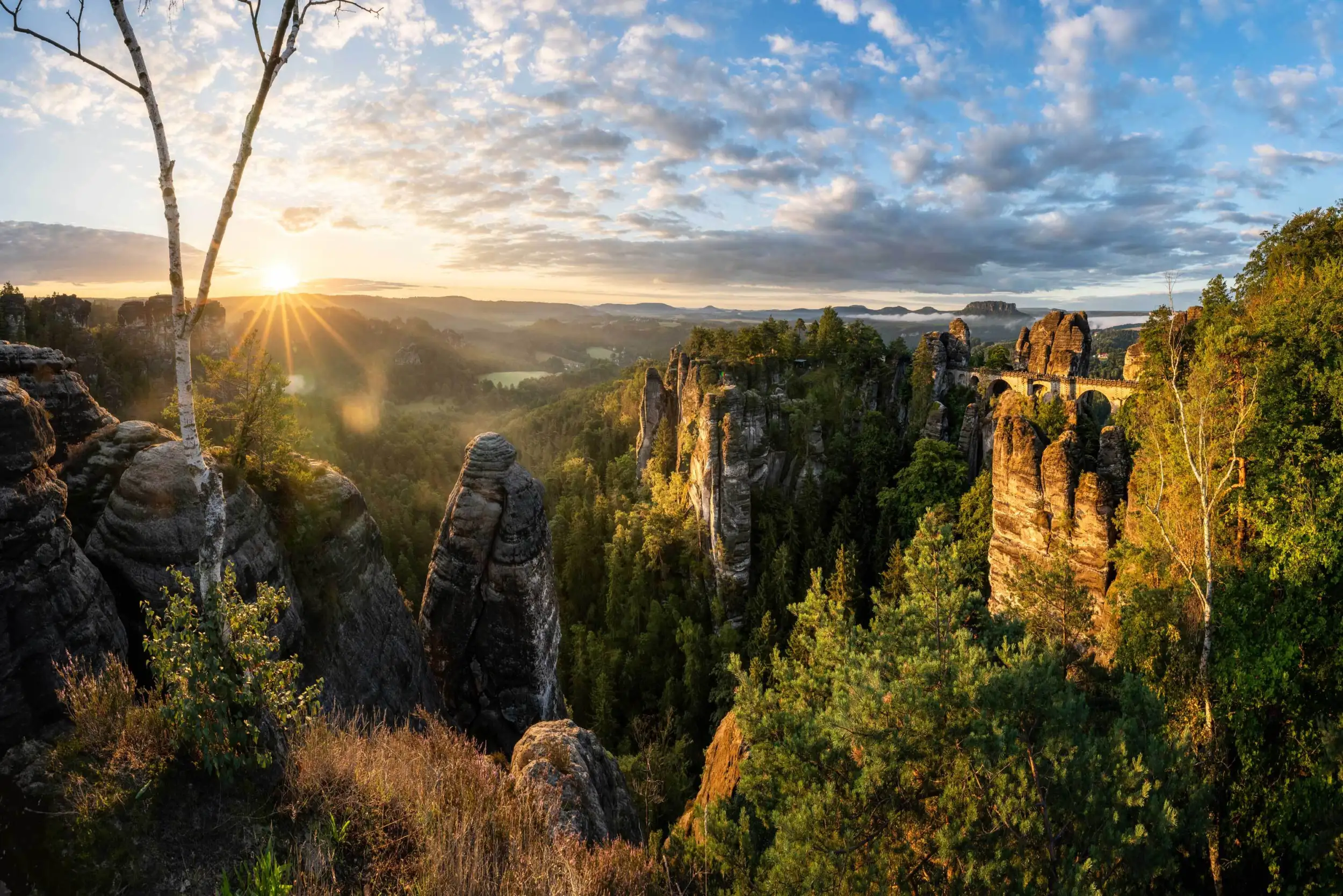 Rock formations and a bridge in a green landscape at sunrise, with a clear sky and scattered clouds.
