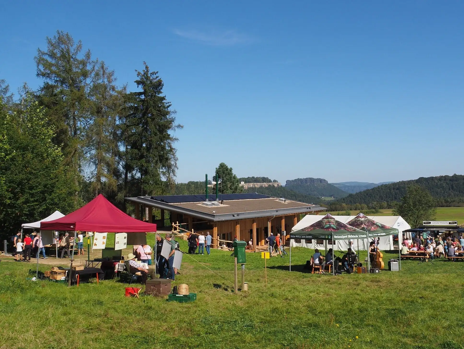 Das Waldfest in Leupoldishain mit Panoramablick zur Festung Lilienstein; auf der Wiese sind viele Stände aufgebaut und Menschen flanieren zu einem Imker