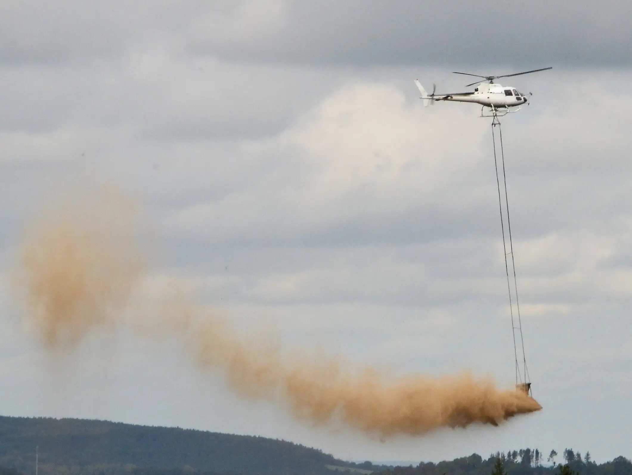 Ein Hubschrauber fliegt über waldige Hügel und lässt gemahlenes Dolomitgestein zur Waldkalkung gezielt ab