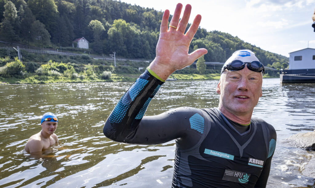 1080 km in ca. 23 Tagesetappen à 46 Kilometer durchschwimmen: Prof Fath richtet mit seinem Extremschwimmen die Aufmerksamkeit auf die Elbe. Als Chemieprofessor engagiert er sich mit der Aktion „Pure Elbe“ für eine plastikfreie Natur zur Verbesserung der Gewässergüte. Am 22. August 2024 gegen 15:50 Uhr erreichte der Professor Postelwitz (Bad Schandau) und Anton Reinhardt (DLRG Ortsgruppe Heidenau) stieg als Schüler der Nationalparkschule dort mit in die Fluten und schwamm mit dem Prof bis Bad Schandau Elbkai. Hier empfangen sie Neuntklässler der Nationalparkschule Königstein, die sich im Rahmen des Projektes „Pure Elbe“ mit Gewässergüte und Mikroplastik befassen. Die Nationalpark- und Forstverwaltung ist als Mitglied von ELBE PARKS – das Netzwerk der Schutzgebiete entlang der Elbe – Teil des Projektes „Pure Elbe“. 
Foto: © Marko Förster